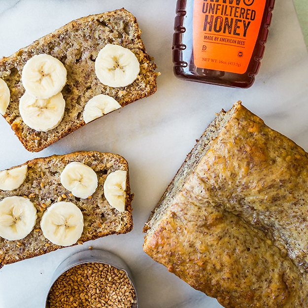 Banana bread with slices cut and topped with banana slices with Nate's honey bottle on a light marble surface.