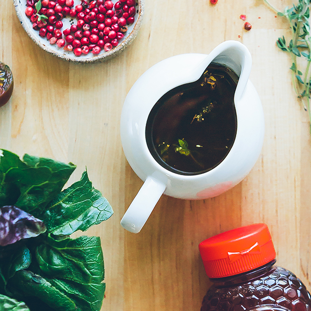 Small white pitcher filled with honey salad dressing, Nate's honey bottle, and fresh herbs on a wooden board.