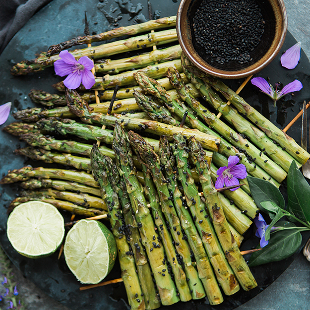 Grilled asparagus on skewers with sesame seeds, lime, and purple flowers on a dark background.