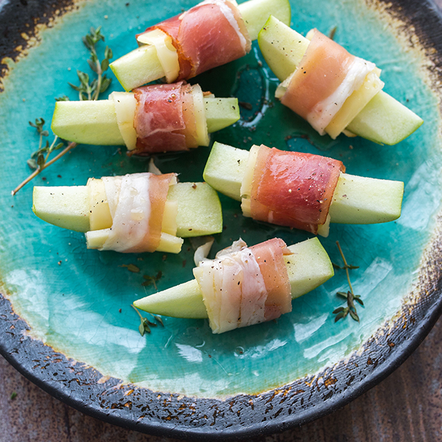 Prosciutto-wrapped melon pieces with cheese, drizzled with honey on a turquoise plate with thyme.