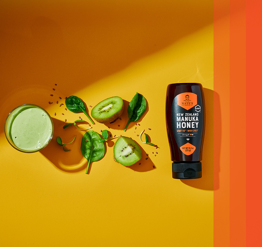 Nate's New Zealand Manuka Honey bottle with a green smoothie, kiwi slices, and spinach leaves on a bright background.