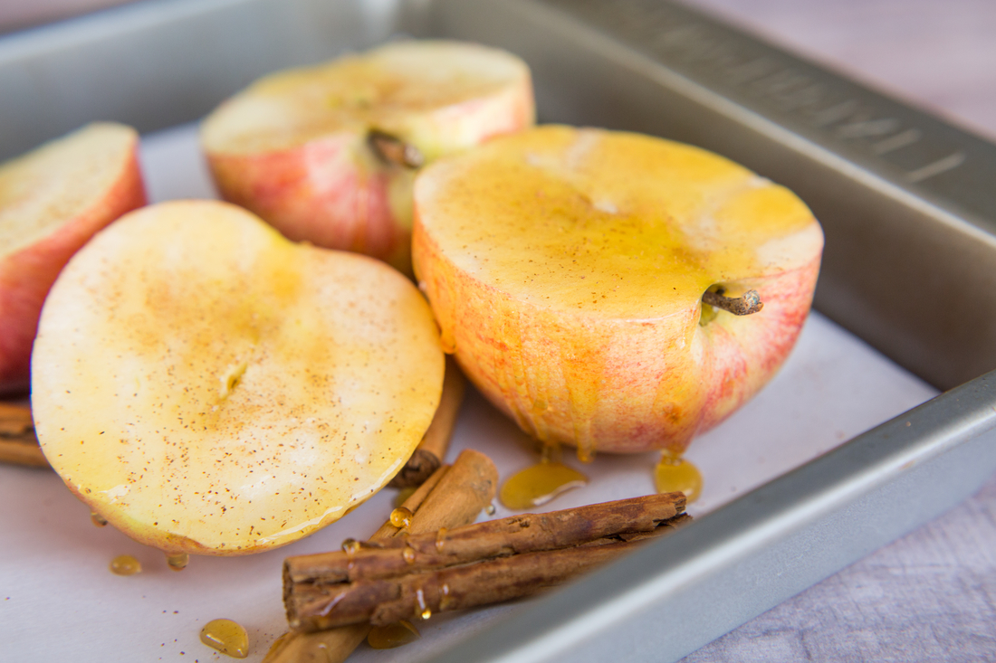 Halved apples sprinkled with cinnamon and drizzled with honey next to cinnamon sticks on baking sheet.
