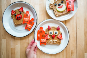 Hand reaching for a plate with nut butter toast decorated with animal faces, with Nate's honey minis on a wooden table.