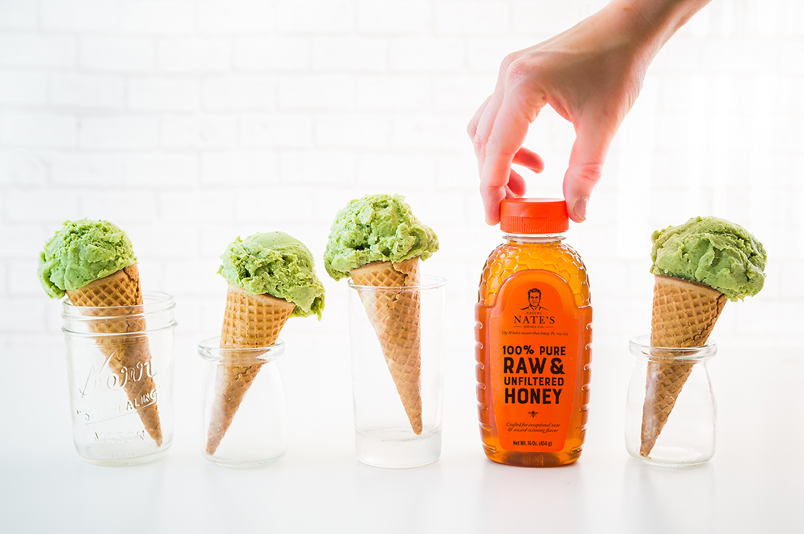 A hand reaches for a bottle of Nate's raw and unfiltered honey, setup of avocado ice cream cones in glasses.