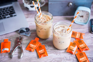 Iced lattes with straws and Nate's honey minis on a desk with a laptop and office tools.
