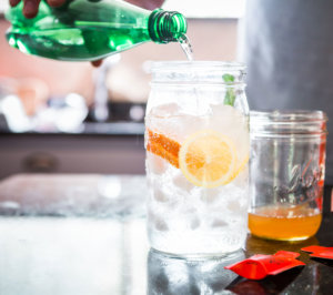 Pouring a drink into a glass with ice and citrus, with Nate's honey minis and a blurred kitchen background.