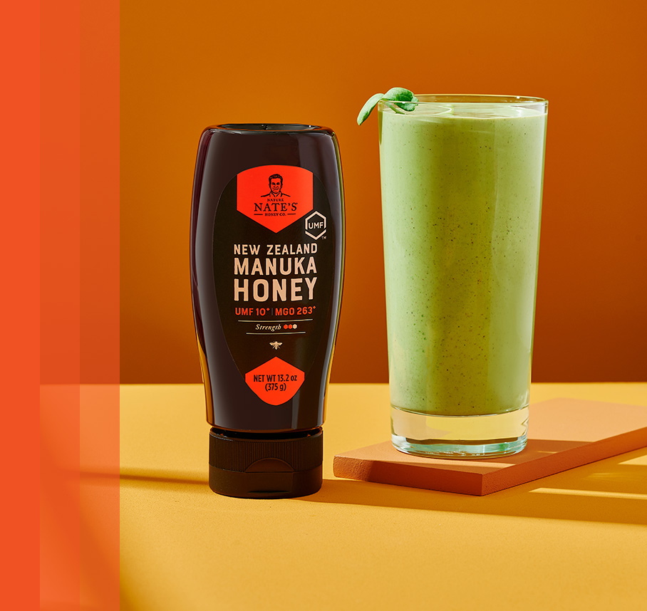 Nate's New Zealand Manuka Honey bottle beside a green smoothie on a yellow surface with an orange background.