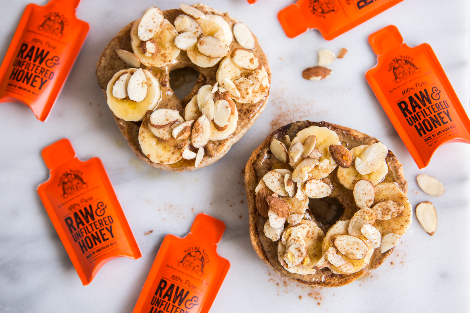 Bagels topped with almond butter, bananas and almonds, with Nate's honey minis scattered on a marble surface.