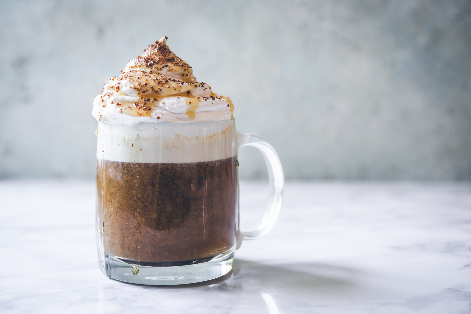 Frothy pumpkin latte with whipped cream topping in a clear mug on marble surface, light textured backdrop.