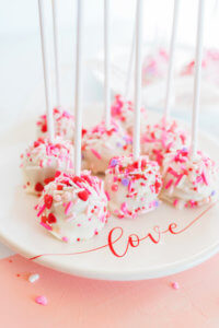 Cake pops with white frosting and pink sprinkles on a plate with "love" written on it.