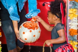 A child in a ladybug costume reaching for a Nate's honey mini from a white pumpkin with orange spots, held by an adult. A child in a ladybug costume reaching for a Nate's honey mini from a white pumpkin with orange spots, held by an adult.