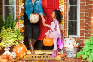 A child dressed in a superhero costume goes trick-or-treating, receiving candy from a person at a decorated front porch.