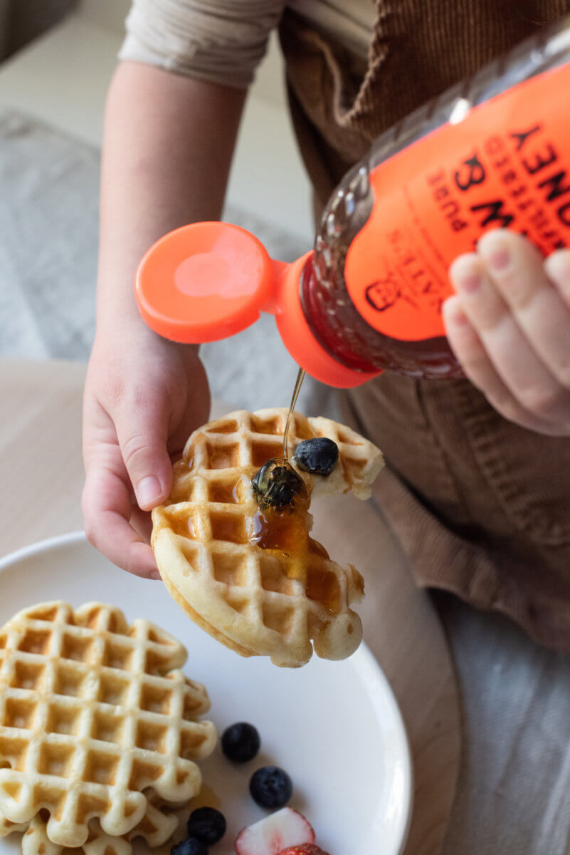 A child's hands pouring Nate’s honey on a waffle with blueberries atop, with more waffles and blueberries on the plate.