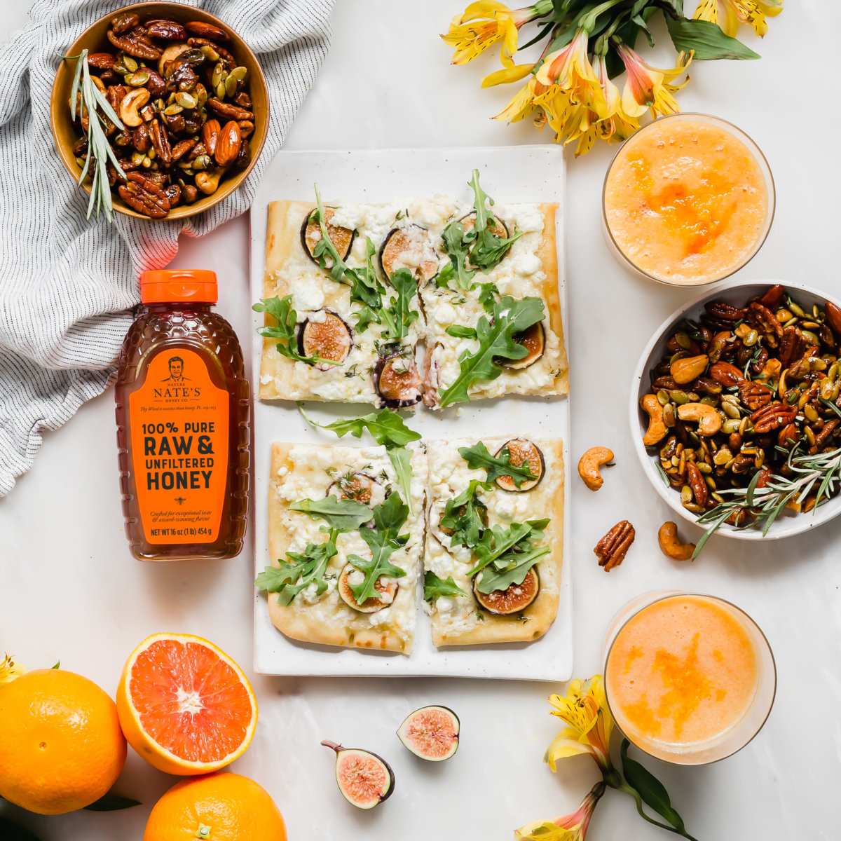 Flatbreads with arugula and figs, a bottle of Nate's honey, bowls of nuts, grapefruits and smoothies on a white surface.