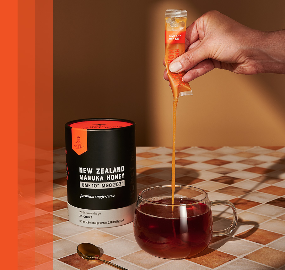 Hand squeezing a Manuka Honey Mini stick into a glass cup of tea, with product container on a tiled table.