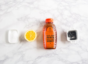 Baking soda in small dish, lemon half, bottle of Nate's honey, small bowl with charcoal capsules, on a marble surface.