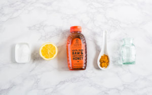 Baking soda in small dish, lemon half, Nate's honey bottle, turmeric, and empty jar on marble surface.
