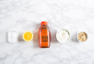 Baking soda in small dish, lemon half, bottle of Nate's honey, cup of yogurt, small bowl of oats, on a marble surface.