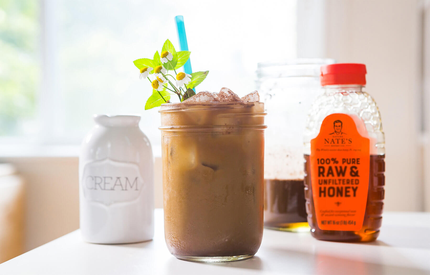 Iced coffee with mint, cream pitcher and a bottle of Nate's honey on a bright table.