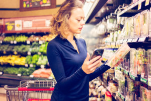 A person in a blue top is reading a label on a product in a supermarket aisle with a shopping cart nearby. A person in a blue top is reading a label on a product in a supermarket aisle with a shopping cart nearby.