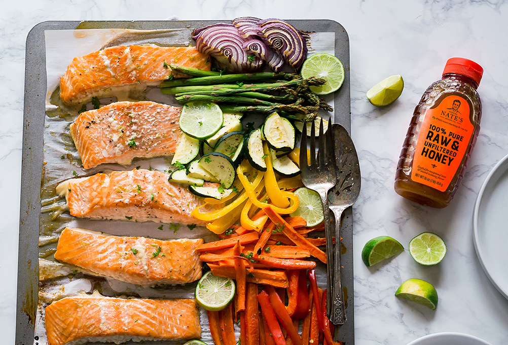 Grilled salmon and vegetables on a sheetpan with a bottle of Nate’s honey and lime wedges on a marble countertop.