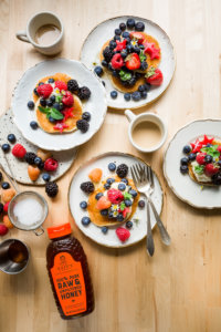 Pancakes with berries on plates, coffee cups, and Nate's honey bottle on a table.