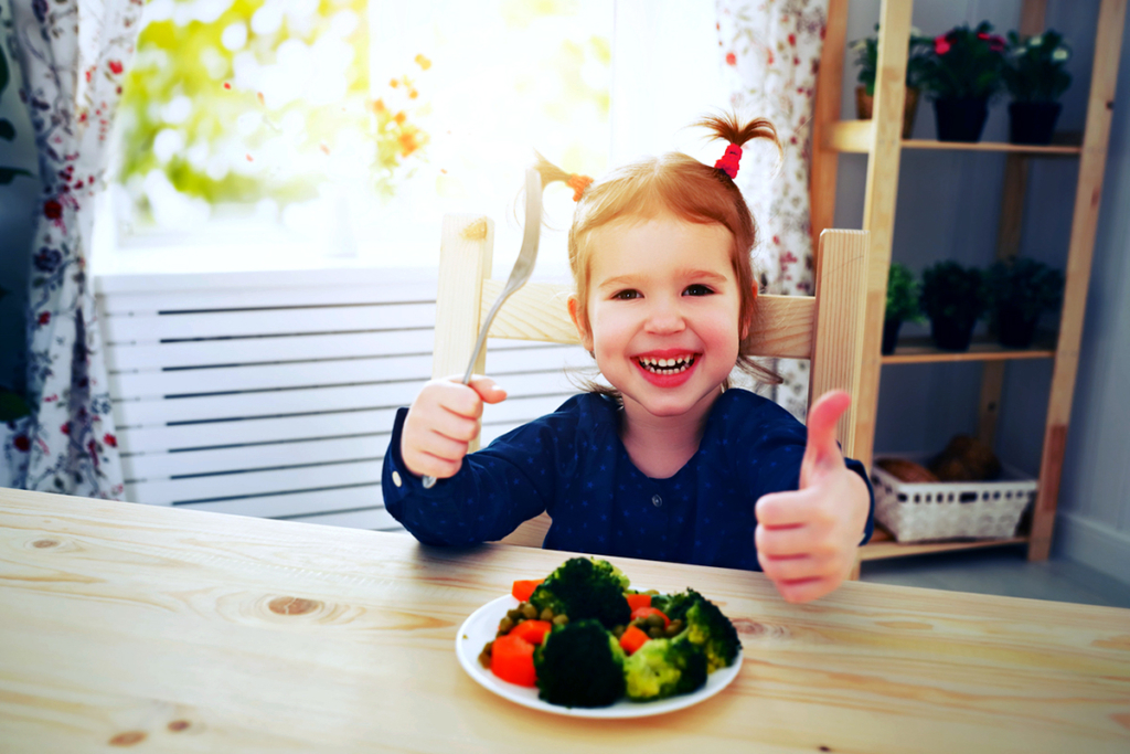 Happy child giving thumbs up to a plate of veggies in a bright room.