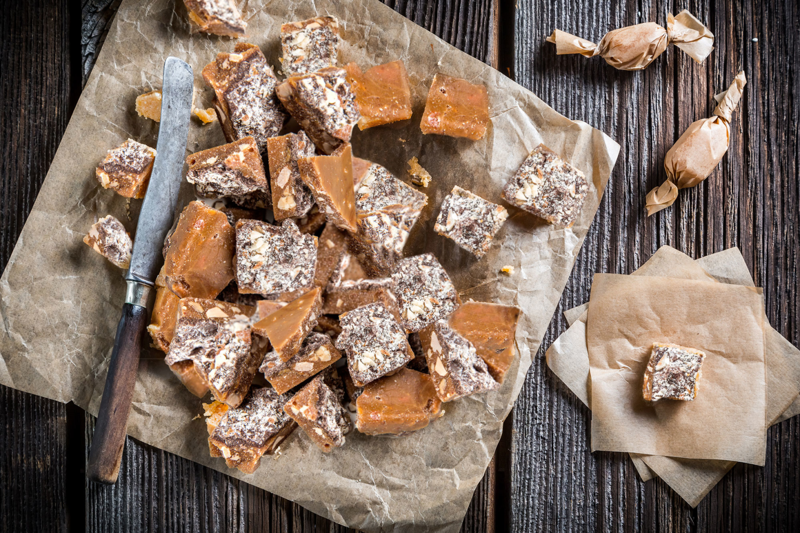 Chunks of golden brown almond roca sprinkled with powdered sugar on crinkled parchment, rustic knife alongside.