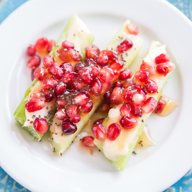 A dish of celery sticks with creamy topping and pomegranate seeds on a white plate, over a blue patterned cloth.