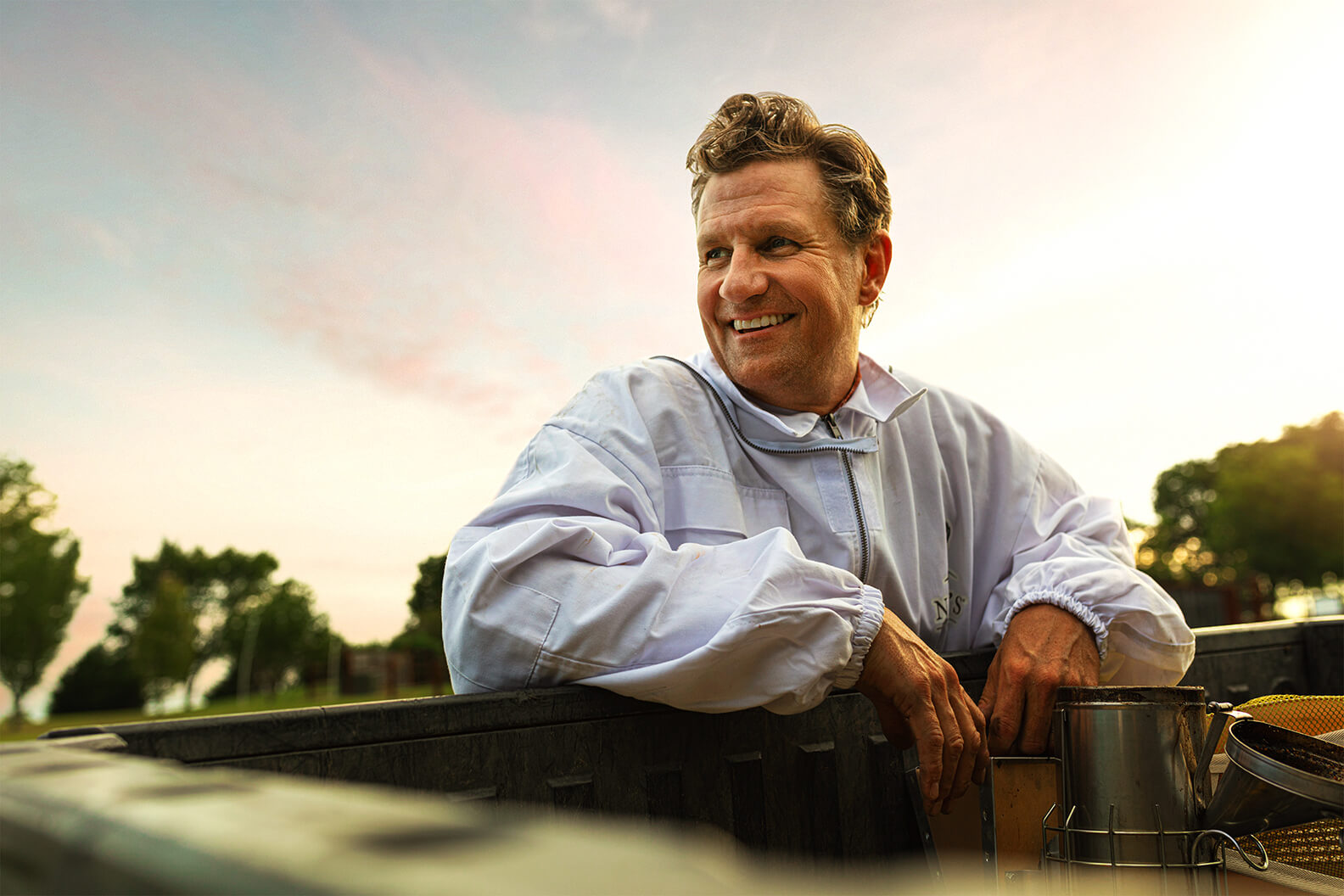 The man Nature Nate smiling, wearing a beekeeping suit and leaning on a honey barrel.