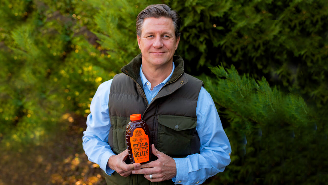 man in a vest holds a bottle of Nate's honey outdoors, with greenery in the background.