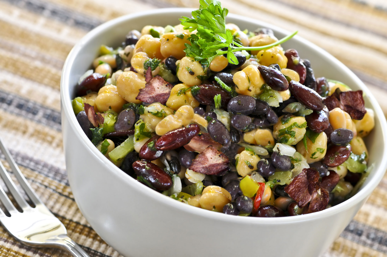 A bean salad with mixed legumes, herbs, and diced vegetables in a white bowl on a plaid cloth with a fork to the side.