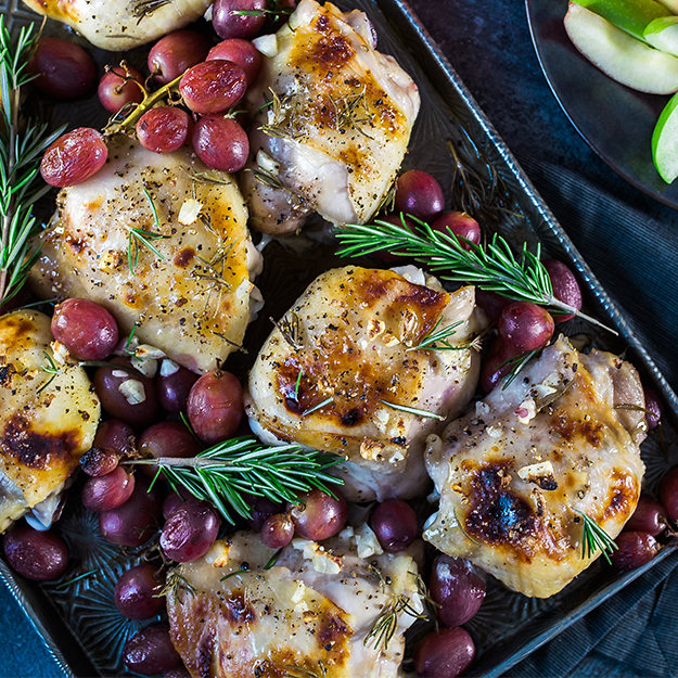 Baked chicken with grapes and rosemary in a pan, on a dark table with green accents.