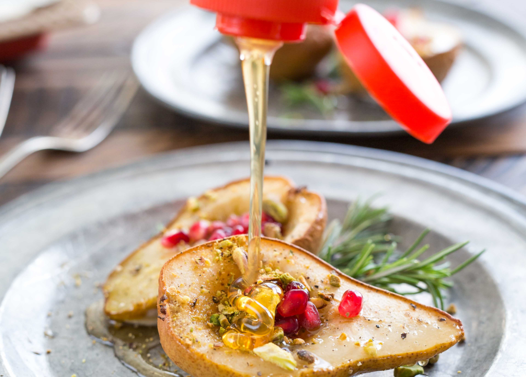 Nate’s honey is being drizzled onto a grilled pear garnished with pomegranate and rosemary on a rustic metal plate.
