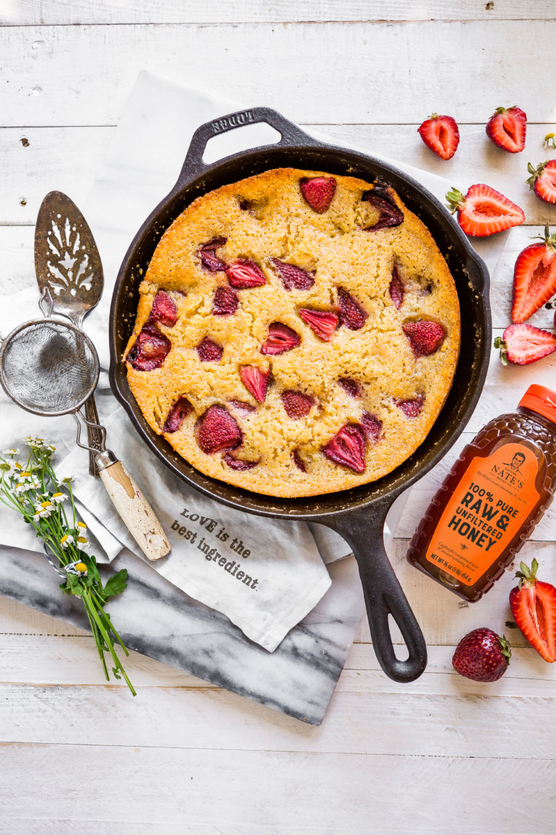 Strawberry cake in skillet, a bottle of Nate’s honey, flowers and silver spoon on a napkin.