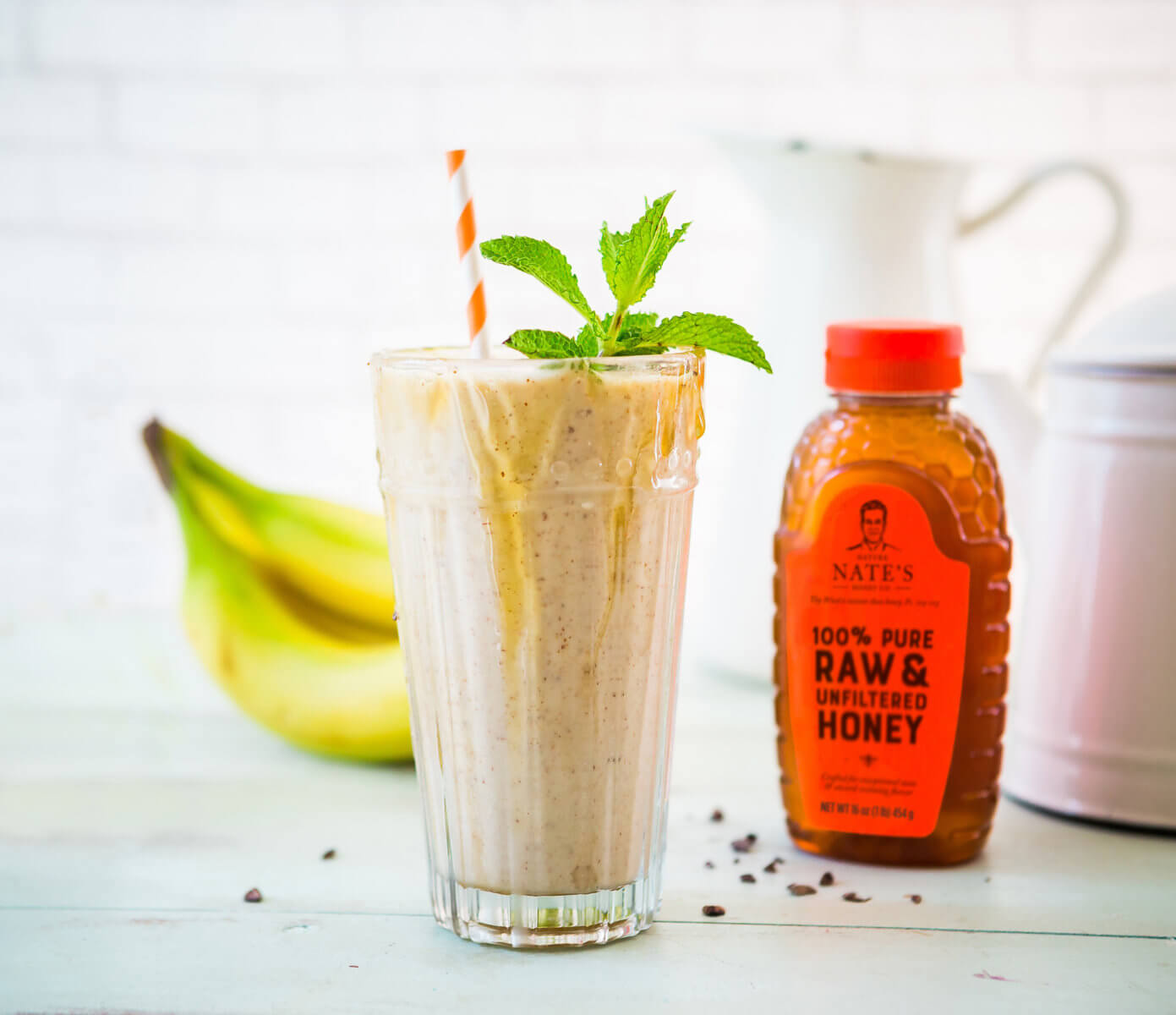 Banana smoothie with mint sprig and honey drizzle, a bottle of Nate's honey, and fresh bananas on a light background.