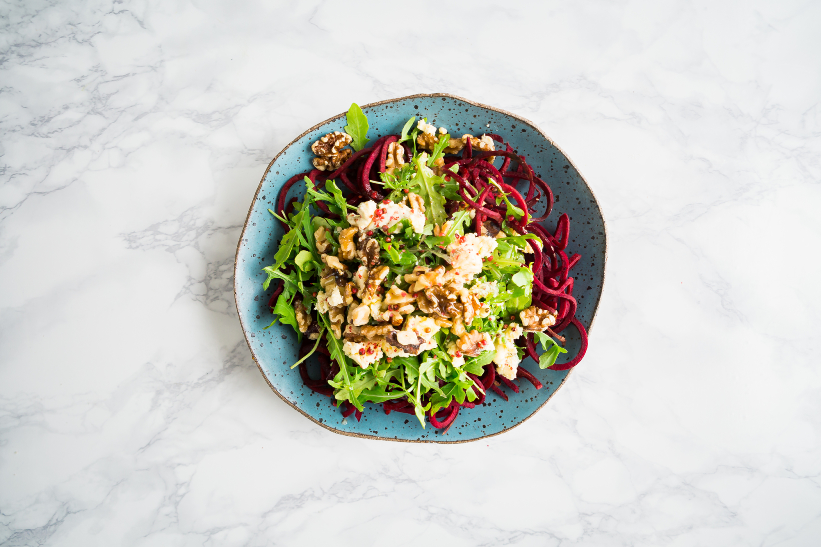 Top-down image of a beet and arugula salad with bleu cheese and walnuts on a blue plate, marble backdrop.
