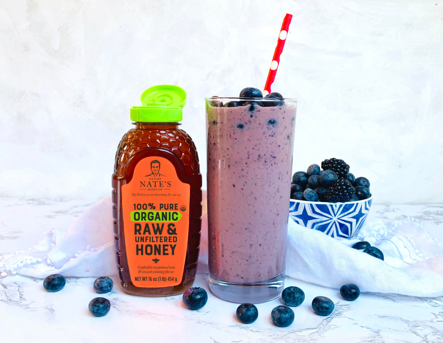 Blueberry smoothie with straw, honey bottle, and bowl of berries on a marbled background.