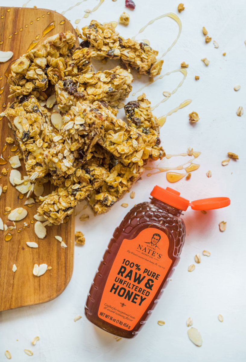 Granola bar pieces on a cutting board with  a bottle of Nate's honey and oats scattered around.