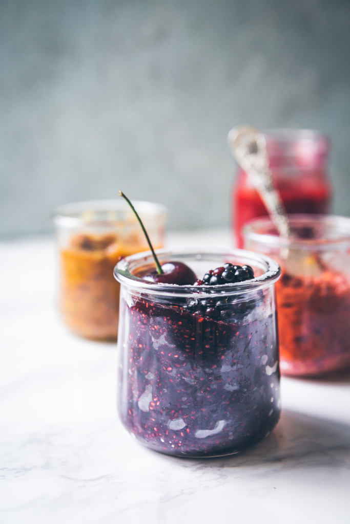 A jar of berry jam with a cherry on top, against a light backdrop.