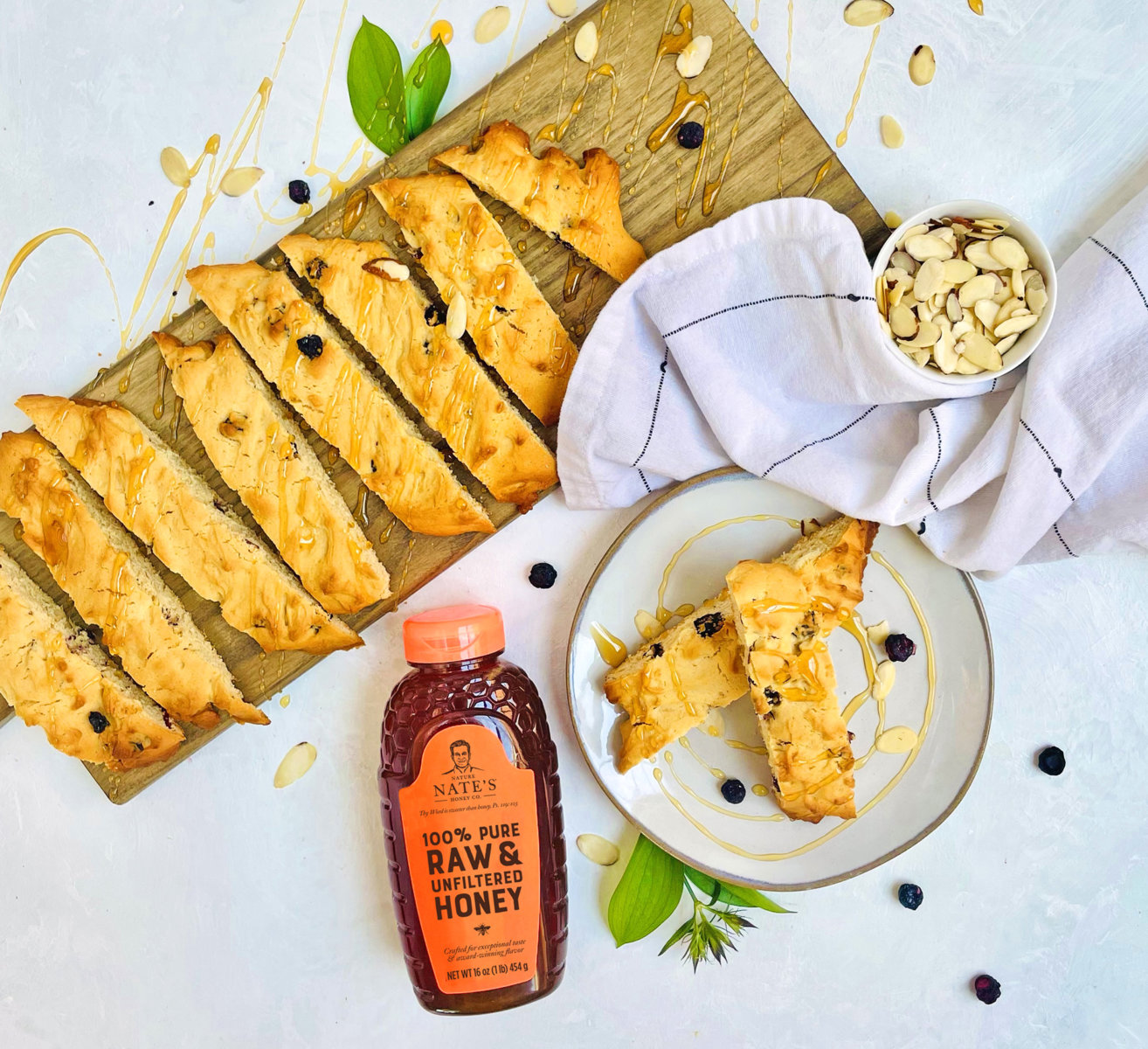 Golden biscotti and almonds on a board, drizzled with honey, with a bottle of Nate’s honey and a blue background.