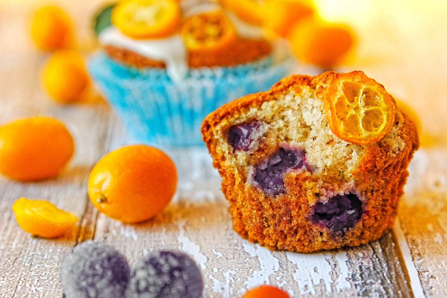 A berry-filled muffin with a kumquat slice on top, surrounded by kumquats and dusted with sugar, on a wooden surface.