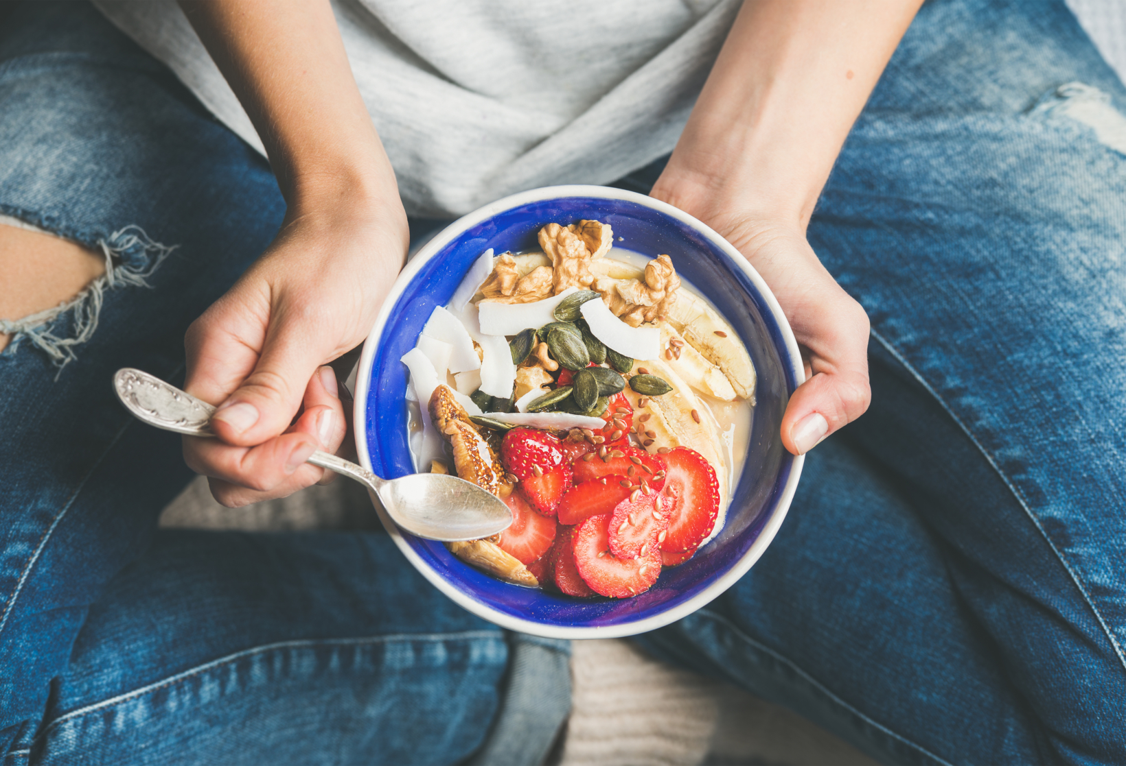 Person in jeans holding a blue bowl of yogurt with strawberries, banana and granola.
