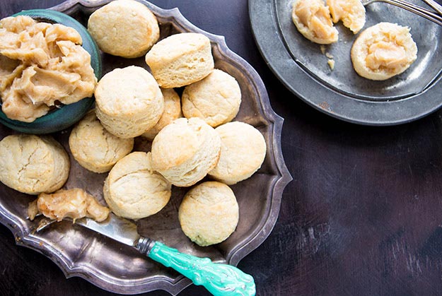 Biscuits on a silver platter, honey butter in a bowl, dark wooden table, teal knife, and dark plate in background.