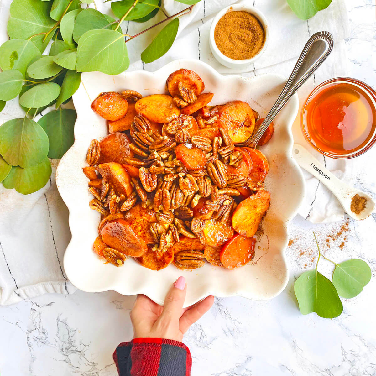 Plate of roasted sweet potatoes and pecans with spices, honey, green leaves, elegantly arranged on a marble background.