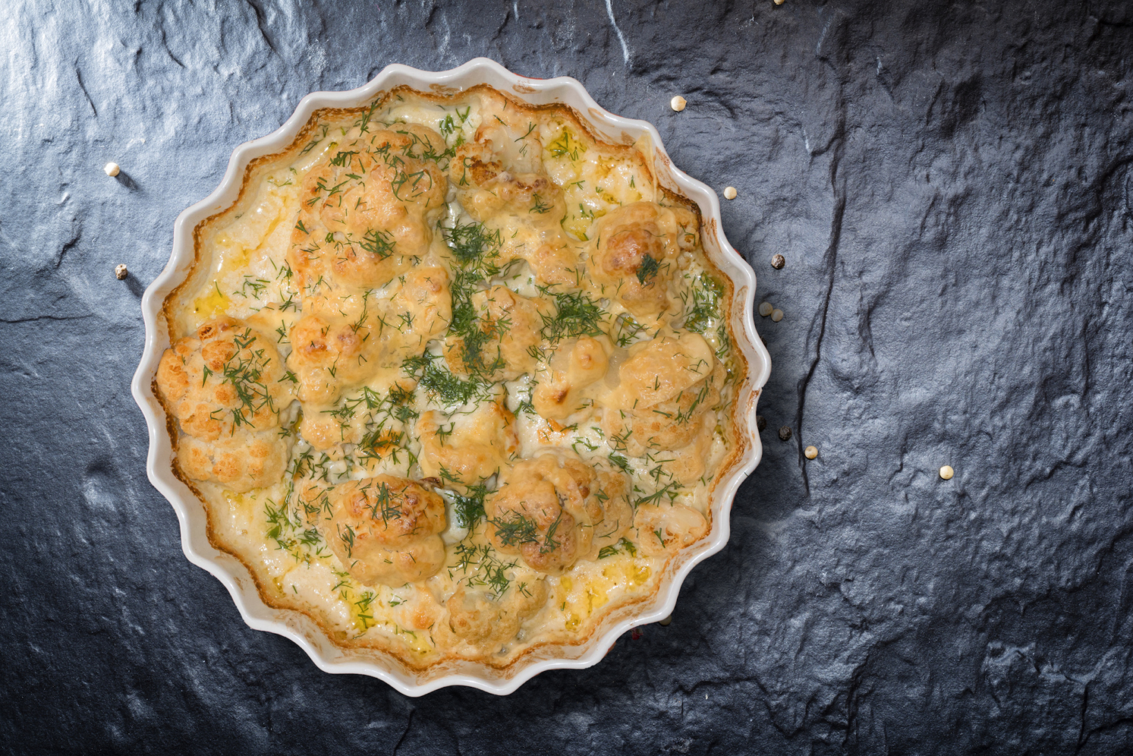 Golden-brown cauliflower grits in a fluted white dish, presented on a slate background with scattered herbs.