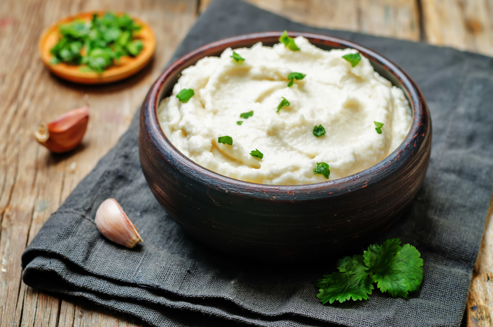 Creamy mashed cauliflower in a dark bowl garnished with green herbs on a gray cloth, garlic cloves, and herb plate.