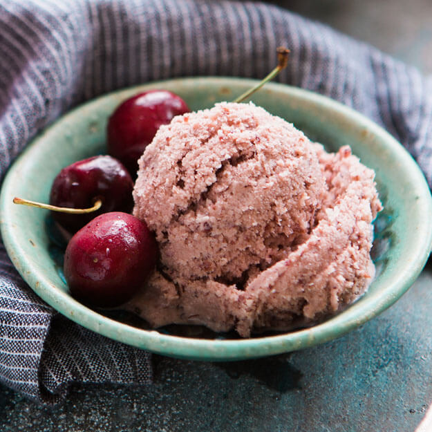 Cherry ice cream in a bowl with cherries, on a blue backdrop with a grey napkin.