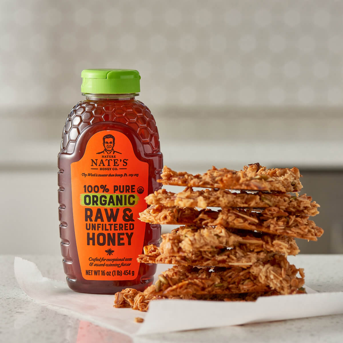 A bottle of Nate’s organic honey next to a stack of granola bark on a marble countertop with a subtle background.