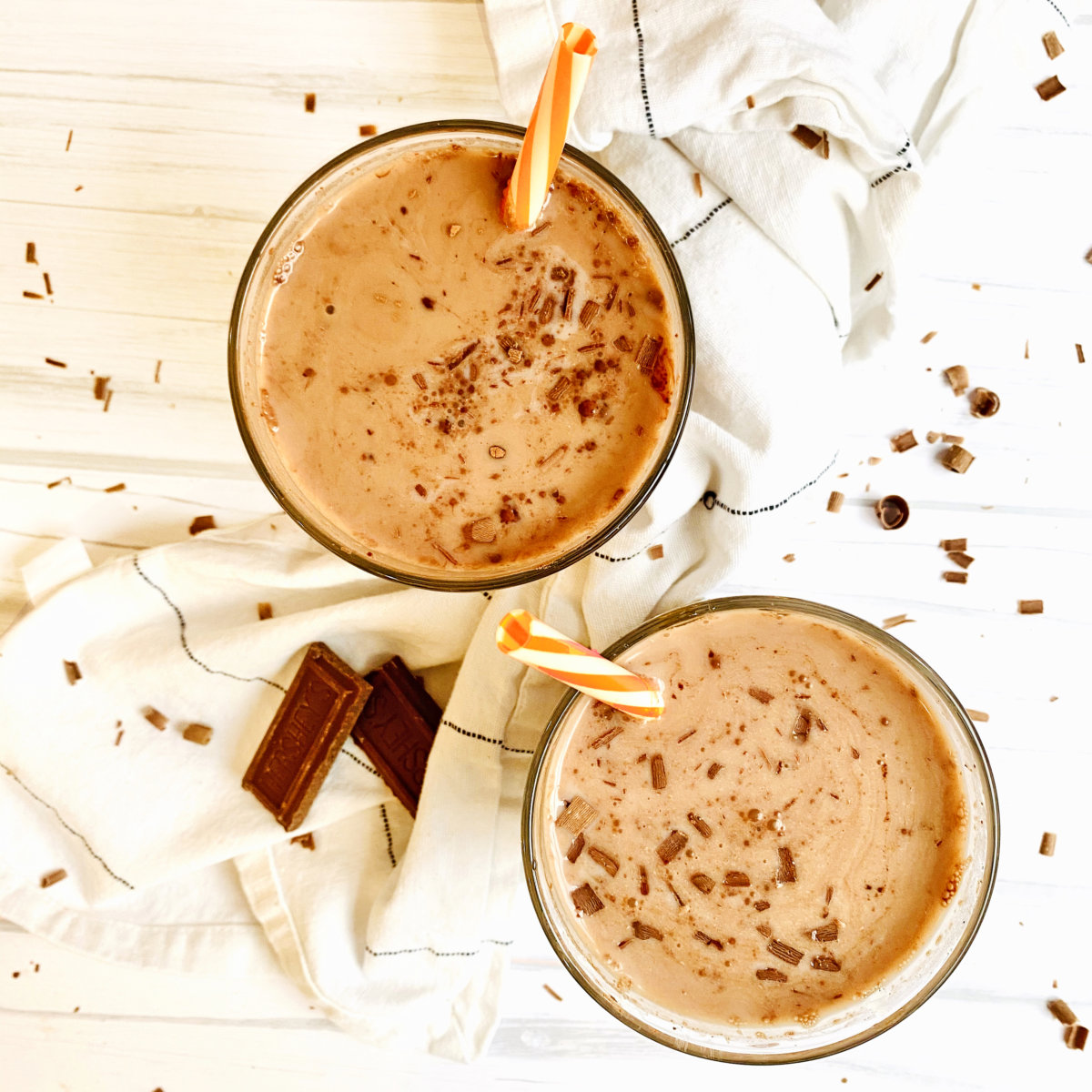Two glasses of creamy chocolate milk, garnished with chocolate shavings, with two small chocolate bars on the side.